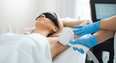 A woman receiving underarm laser hair removal from a blue-gloved technician at a skin and wellness center.