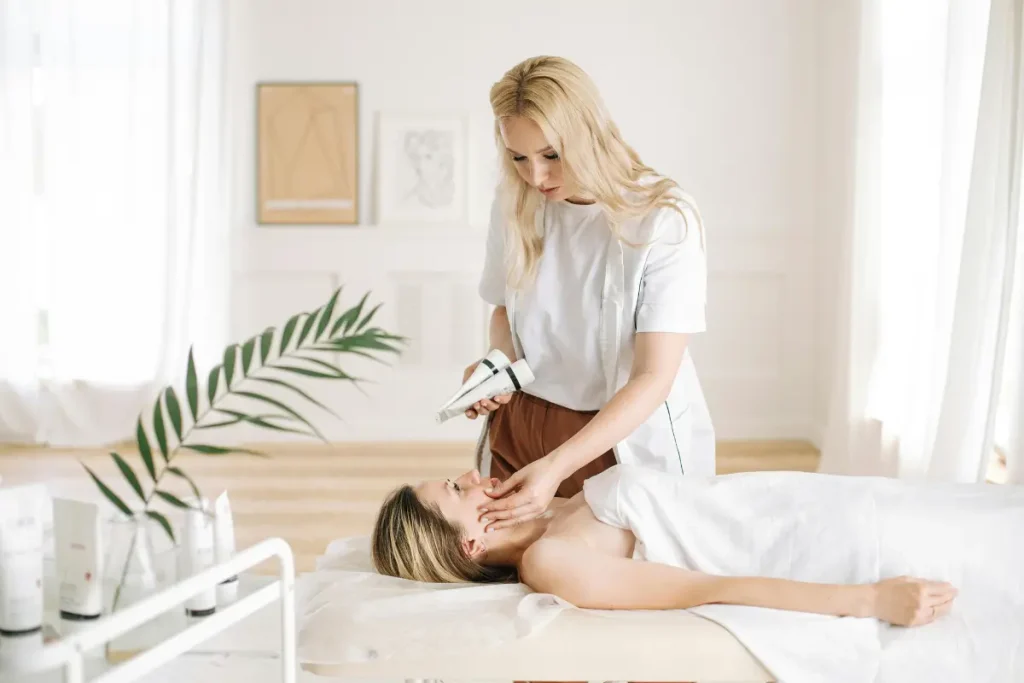 Hydrafacial treatment as an esthetician glides a handheld device across a client's cheek while supporting her face on a treatment bed.