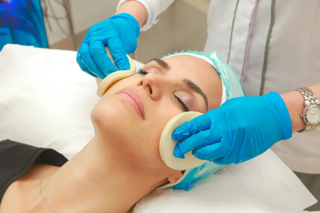 Hydrafacial treatment as a gloved esthetician cleanses a client's face with two soft sponges in a medical spa.