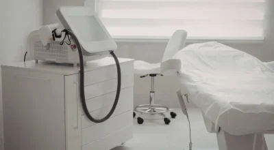 Laser hair removal treatment room with a laser machine, stool, and white treatment bed in a clean, brightly lit medical spa.