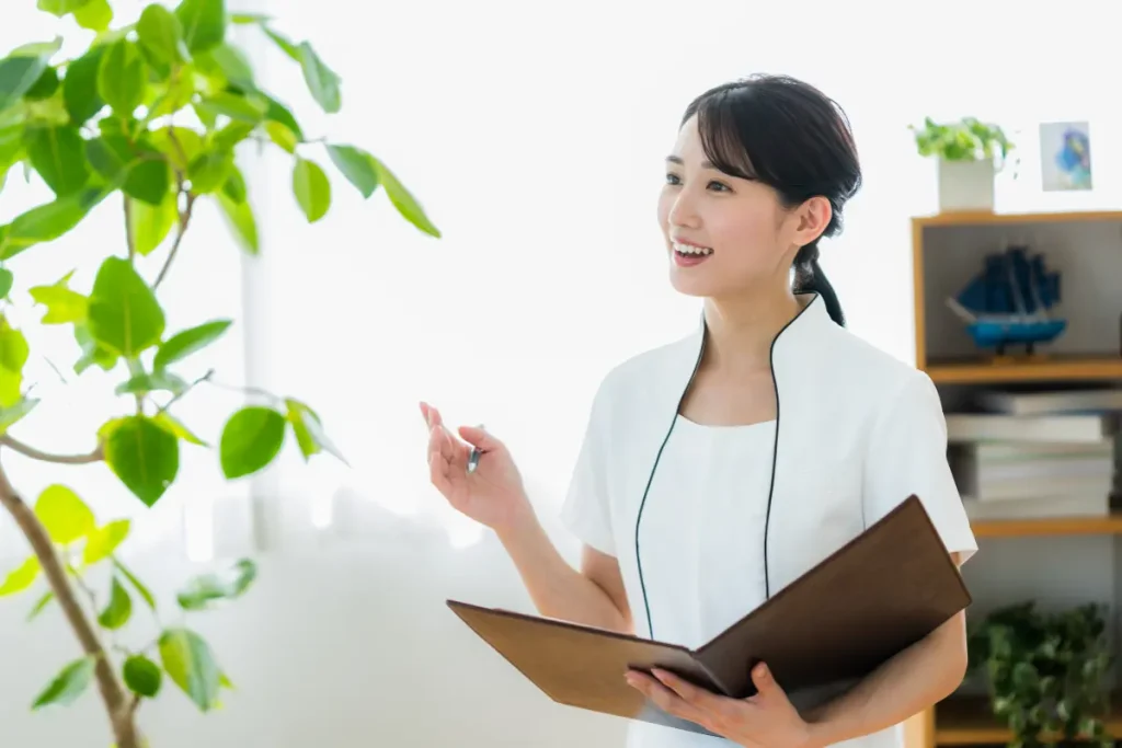Wellness consultant in a white uniform smiling and holding an open patient folder beside a leafy plant in a bright medical spa office.